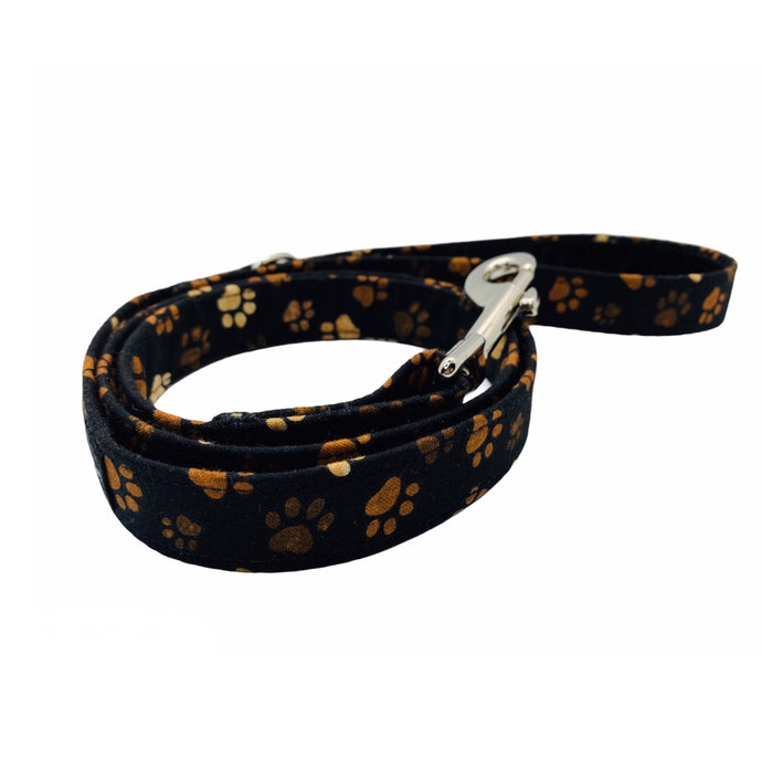 Hand made dog lead in cotton poplin muddy paw print in shades of brown on a black background.  Zinc alloy trigger hook and velvet lined handle. Hand made in the UK and washable.